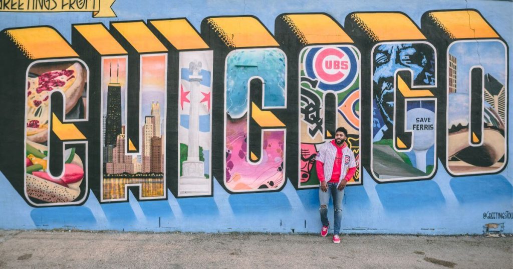 Man wearing jeans red hoodie and sneakers and a cubs baseball shirt leaning against blue wall with street art design stating 'Chicago'
