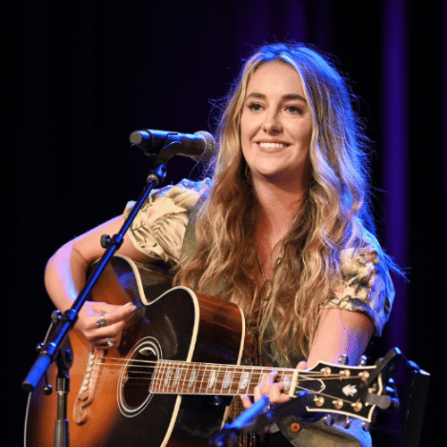 Photo of Lainey Wilson sitting on a stool, playing guitar and smiling. There is a standing microphone in front of her.